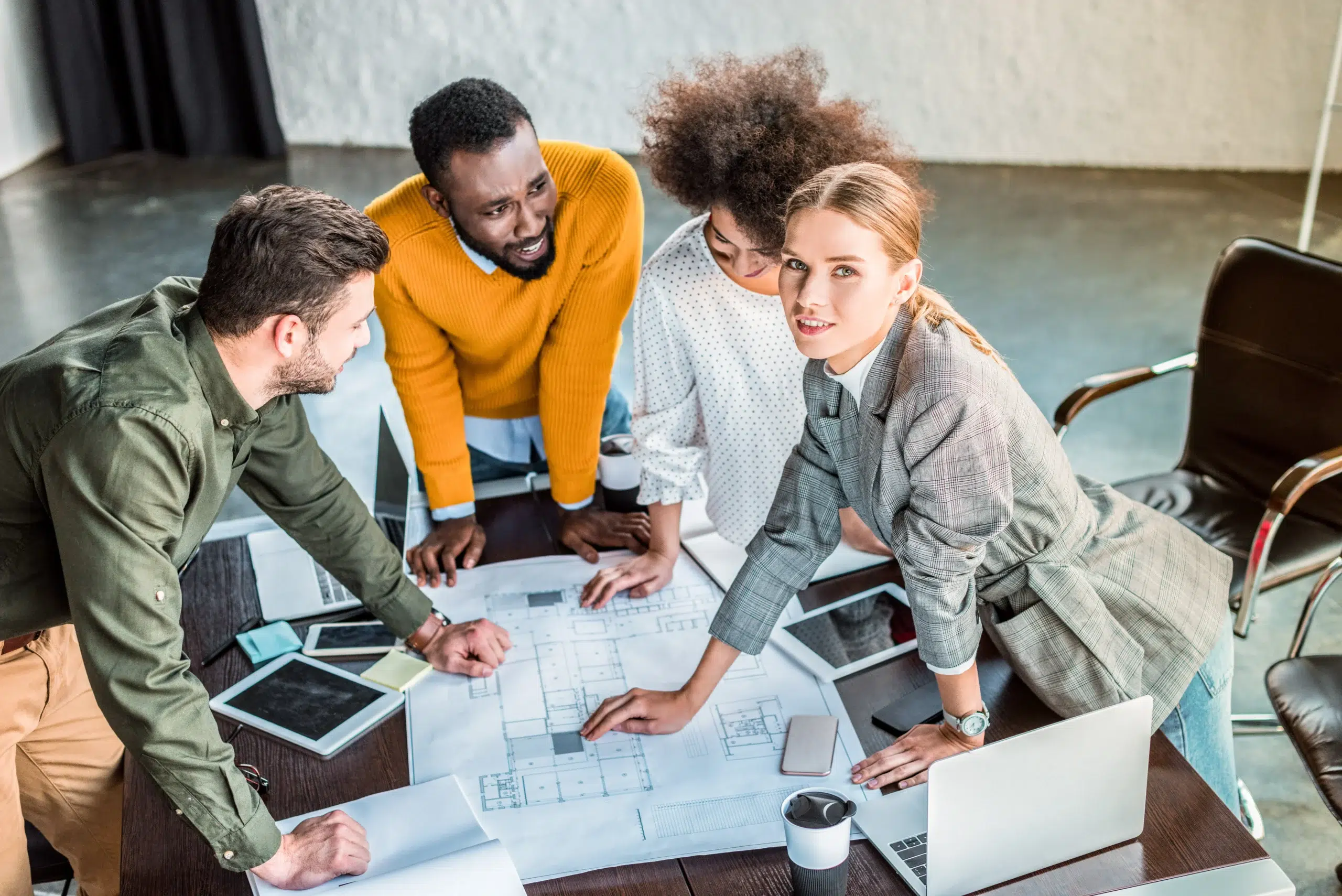 Top-down view of a diverse group discussing complex blueprints spread across a modern wooden table.