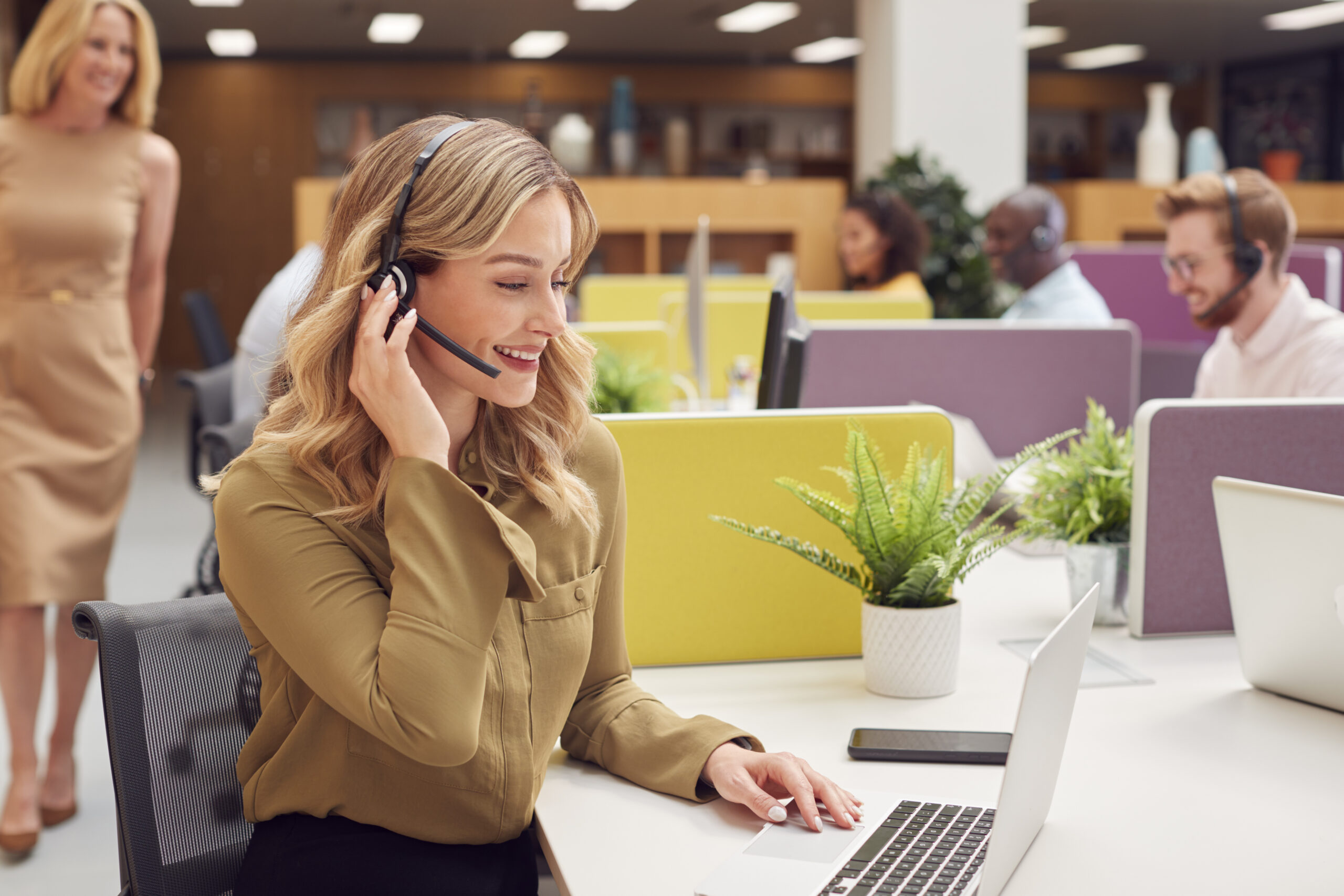 A friendly business woman wears a phone headset while working on a laptop, a professional back office BPO services agent.